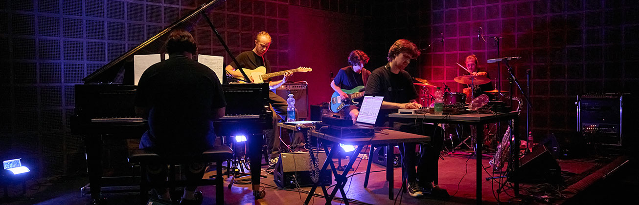 Eine Band mit sechs Musikern spielt auf einer dunkel beleuchteten Bühne, darunter Klavier, Gitarre, Keyboard und Schlagzeug.