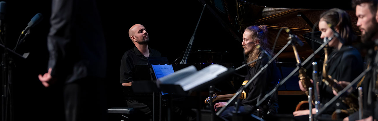 Der Pianist Shai Maestro und weitere Musiker:innen der HSLU Big Band sitzen auf einer Bühne, im Vordergrund steht ein Dirigent.