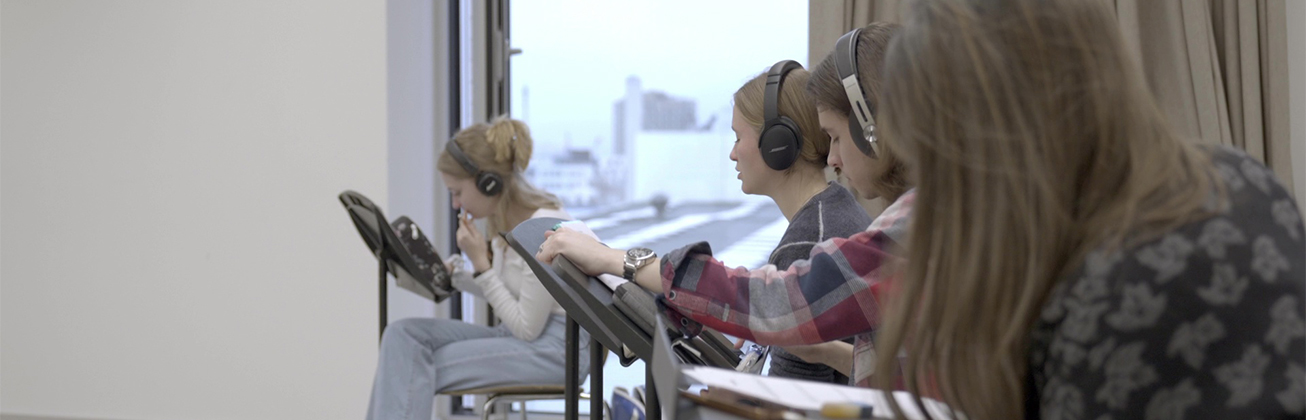 Pre-College-Studierende in einem hellen Unterrichtszimmer, die Noten auf Notenständer studieren und Kopfhörer tragen. 