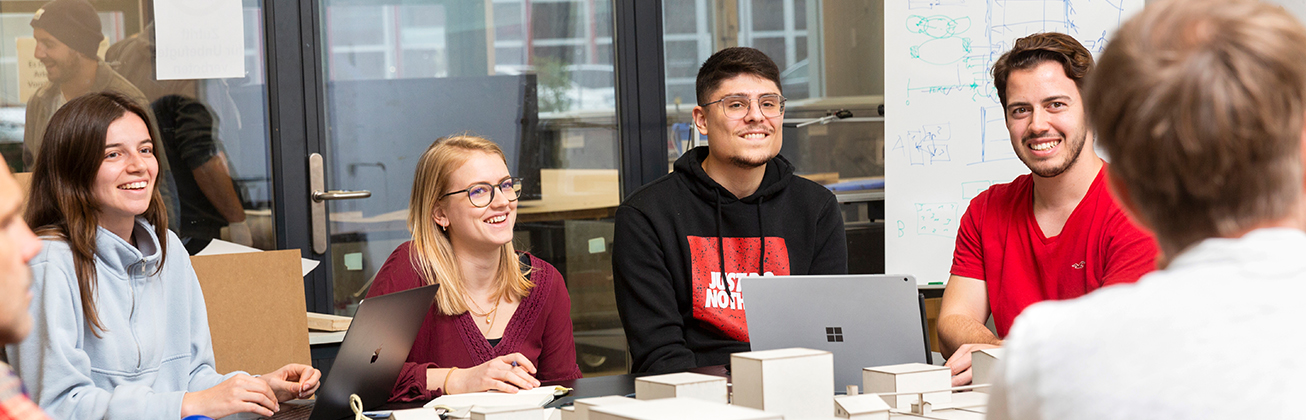 Studierende, die am Tisch mit ihren Laptops sitzen, schauen gespannt und fröhlich in Richtung eines Mannes, der der Kamera den Rücken zukehrt