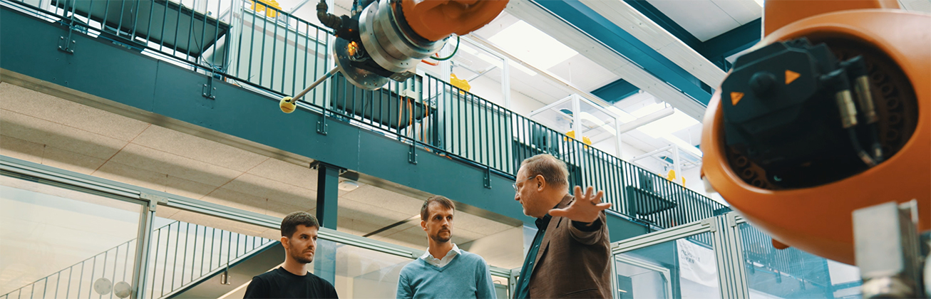 Drei Personen stehen in einer modernen Industriehalle und diskutieren unter einem Industrieroboterarm – Ausbildung und berufliche Qualifizierung im Bereich Robotik und Automatisierung