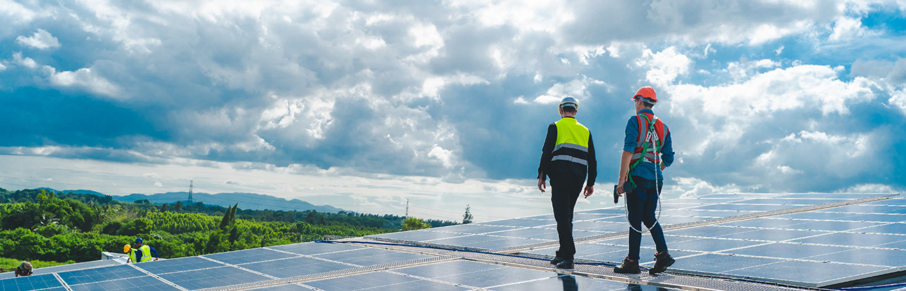 Zwei Techniker mit Sicherheitsausrüstung gehen auf einer großen Solaranlage unter wolkigem Himmel.