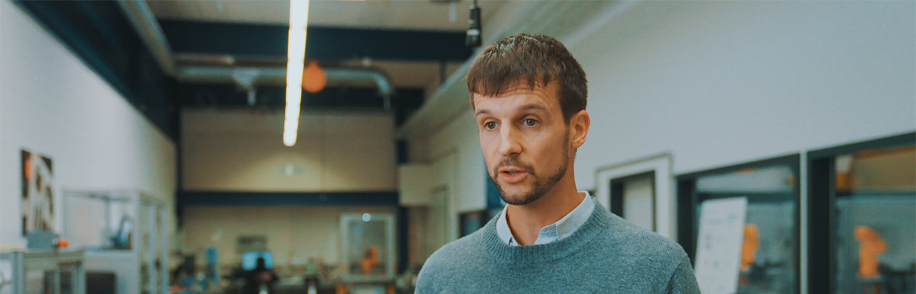 Studiengangleiter Lukas Müller spricht in einer modernen Werkstatt, im Hintergrund sind Arbeitsplätze und industrielle Anlagen zu sehen.