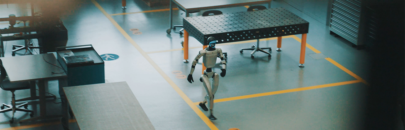 Humanoider Roboter bewegt sich durch eine moderne Forschungs- und Industriehalle mit Werkbänken und technischen Arbeitsplätzen.