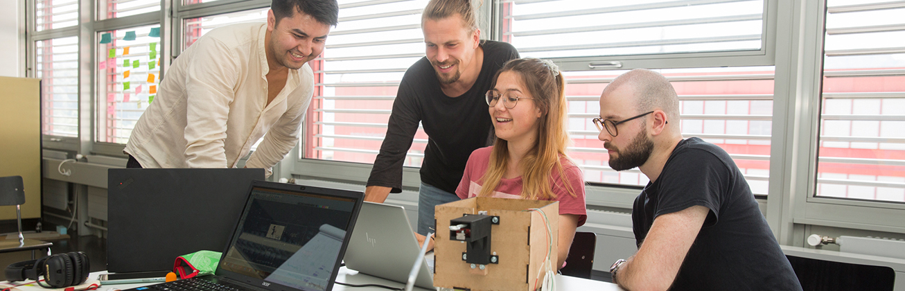 Vier Studierende, die sich zusammen etwas im Computer betrachten und glücklich darüber scheinen