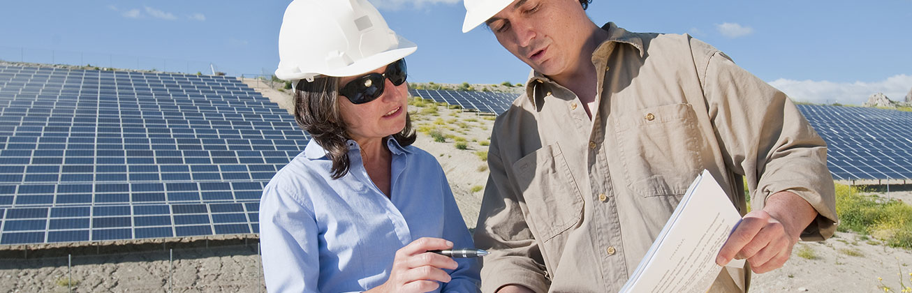 Two engineers wearing hard hats review plans in front of a solar power plant.