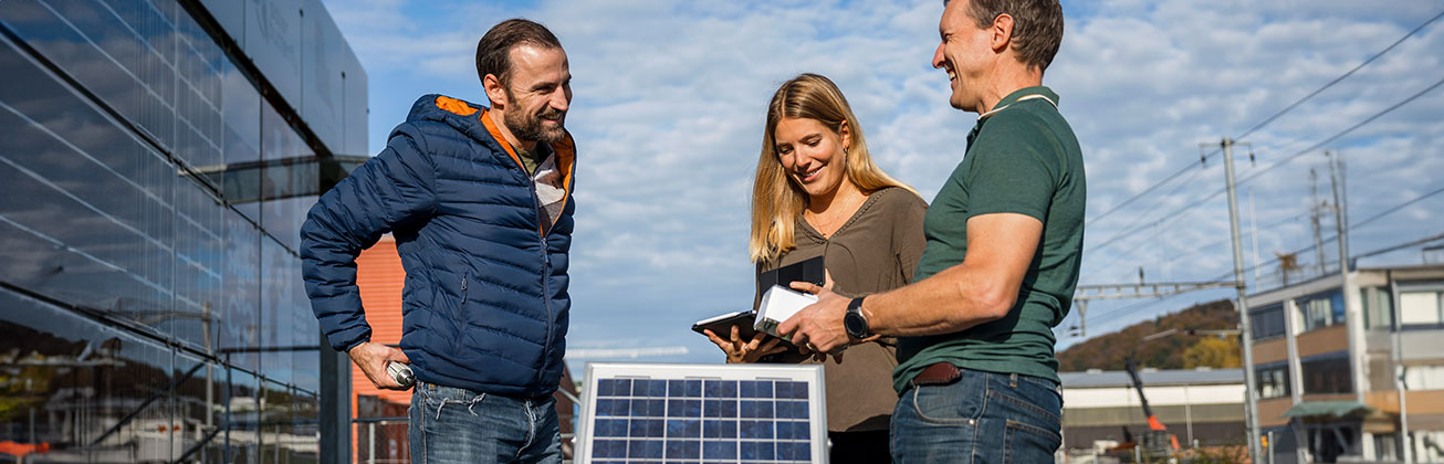 Three people are standing outside around a solar module and are disussing something.
