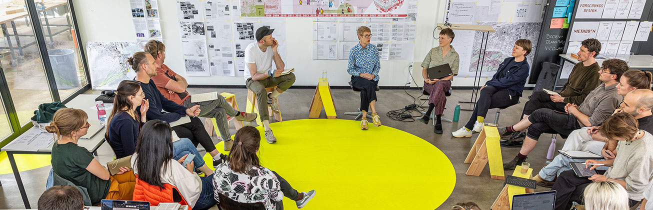 Gruppe von Studierenden sitzt im Kreis in einem hellen Seminarraum mit Plakaten, Notizen und Laptops.