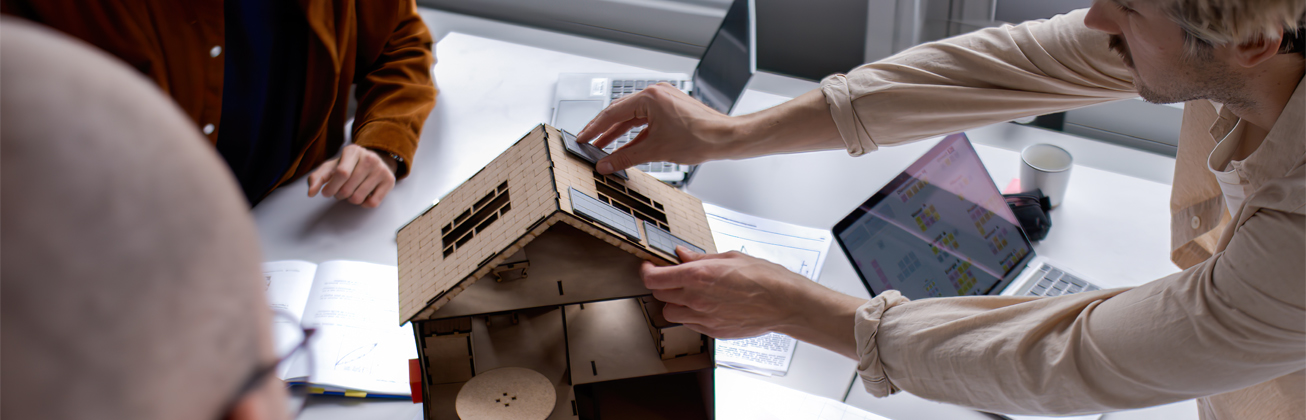 Student fügt ein Teil einem Haus-Modell hinzu, andere Mitsudenten stehen am Rand und schauen zu