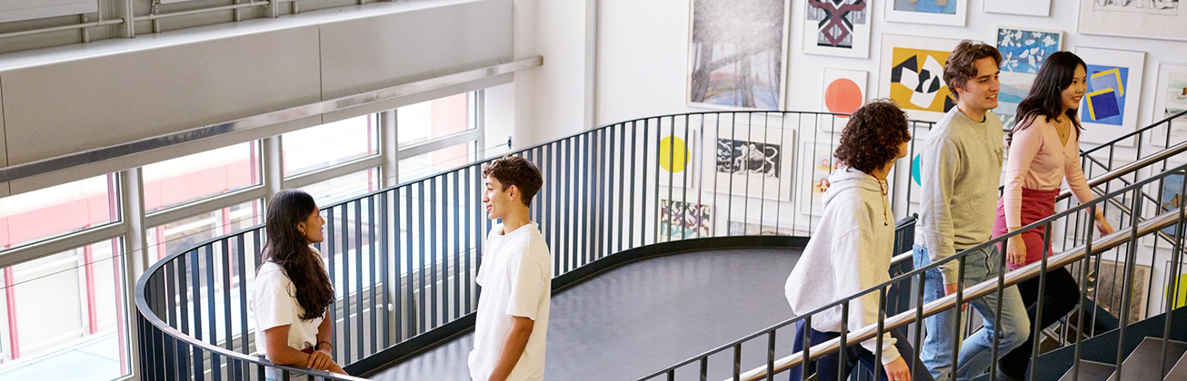 Studierende gehen eine Treppe in einer Hochschule hinauf und unterhalten sich – freundliche Campus-Atmosphäre mit Kunstwerken an den Wänden.