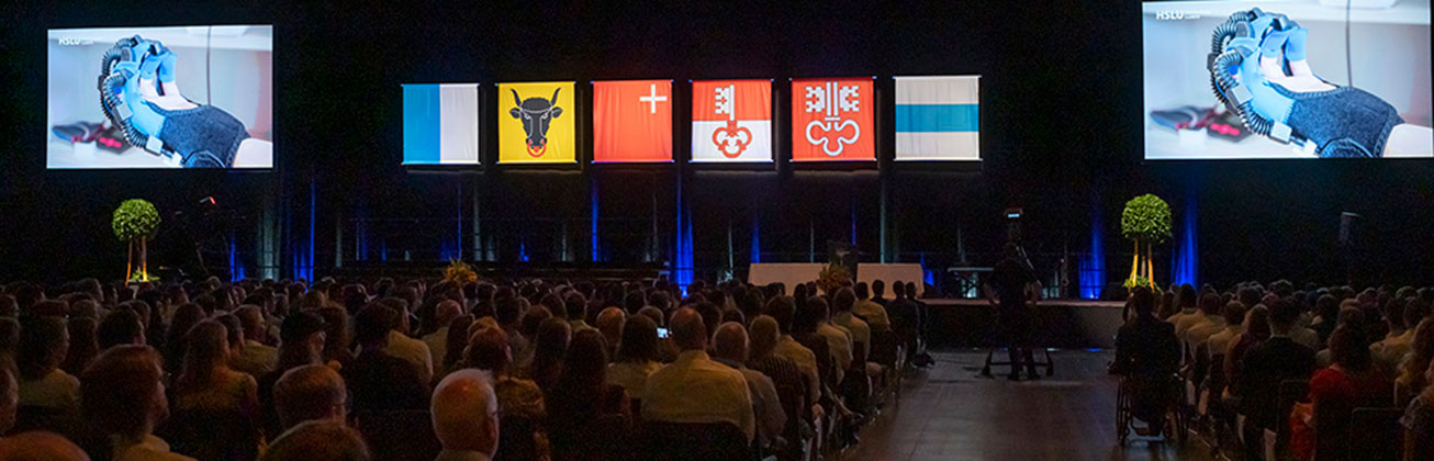 Teilnehmende der Diplomfeier Schauen sich ein Video auf den Bildschirmen der Bühne an.