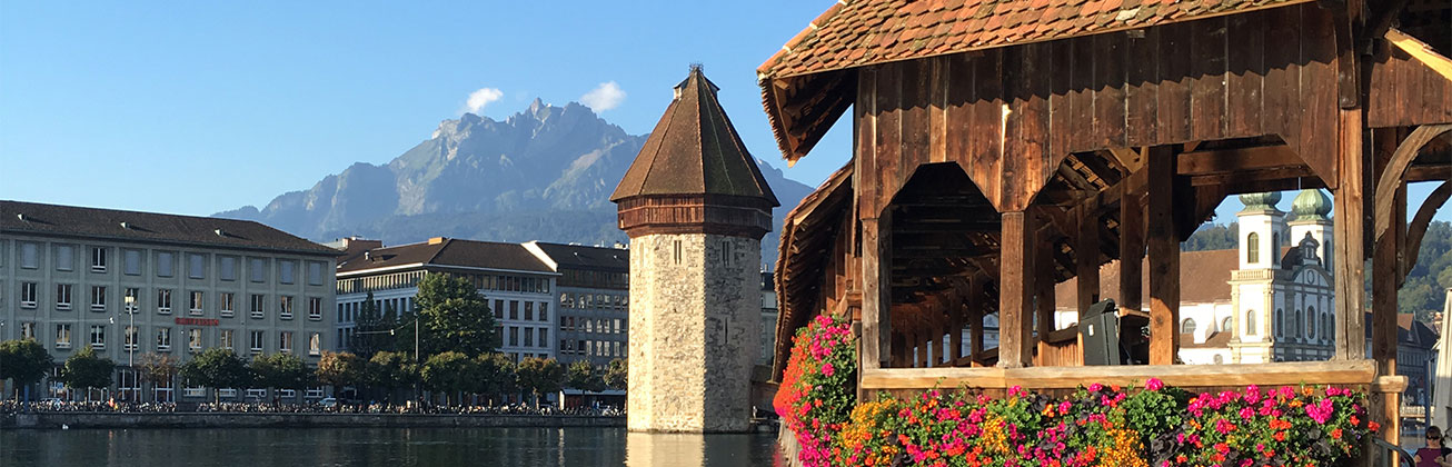 Wasserturm in der Mitte des Bildes, Rechts das Ende der Kapellbrücke, links Gebäude und im Hintergrund blauer Himmel mit dem Pilatus