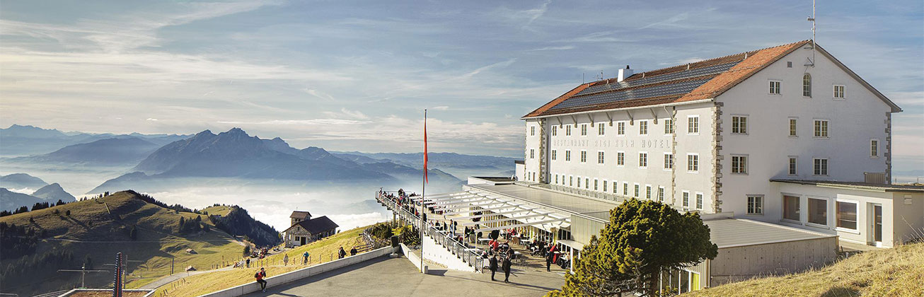 Hotel Rigi auf der rechten Bildseite, leicht bewölkter Himmel, im Hintergrund Berge und grüne Wiesen
