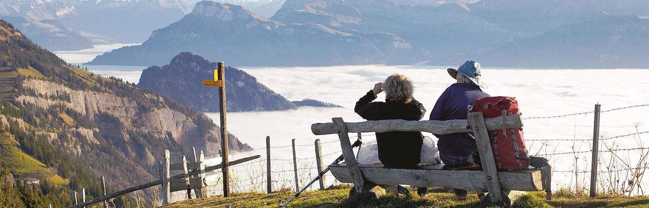 zwei Wanderer sitzen auf einer Holzbank, eine Person hält einen Feldstecher, im Hintergrund Nebelmeer und vereinzelte Berge, die herausragen