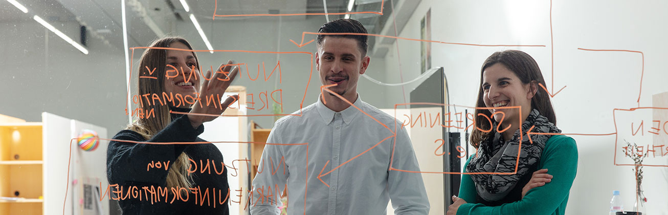 Drei Studierende diskutieren Ideen an einer Glaswand mit handgeschriebenen Notizen.
