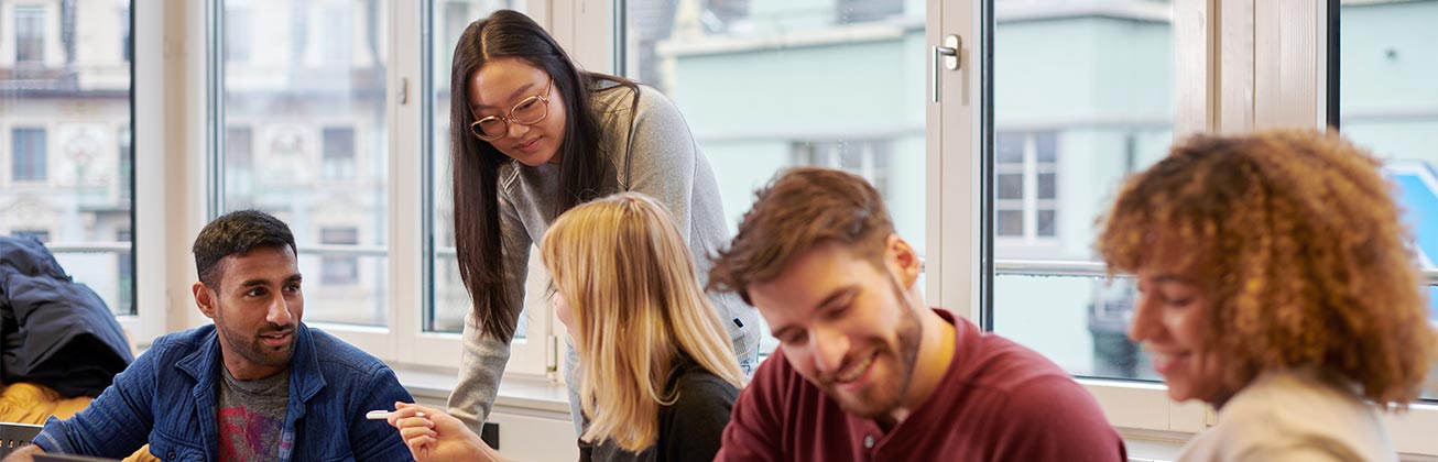 Studierende miteinander im Gespräch am Tisch in einem Unterrichtszimmer und im Hintergrund die Aussicht auf Häuser Luzerns