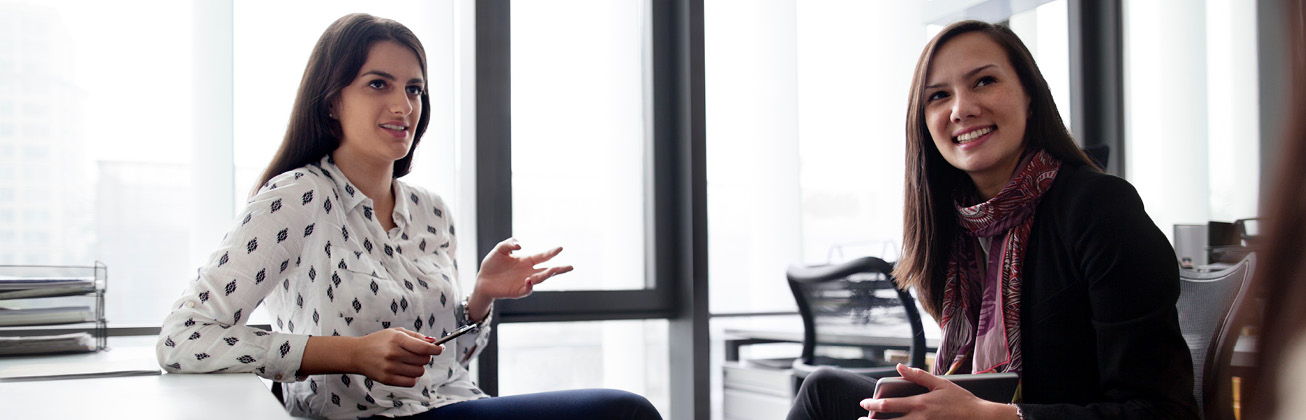 eine Frau mit weisser Bluse mit schwarzen Punkten und eine Frau mit schwarzen Pulli sitzen im Büro und tauschen sich aus