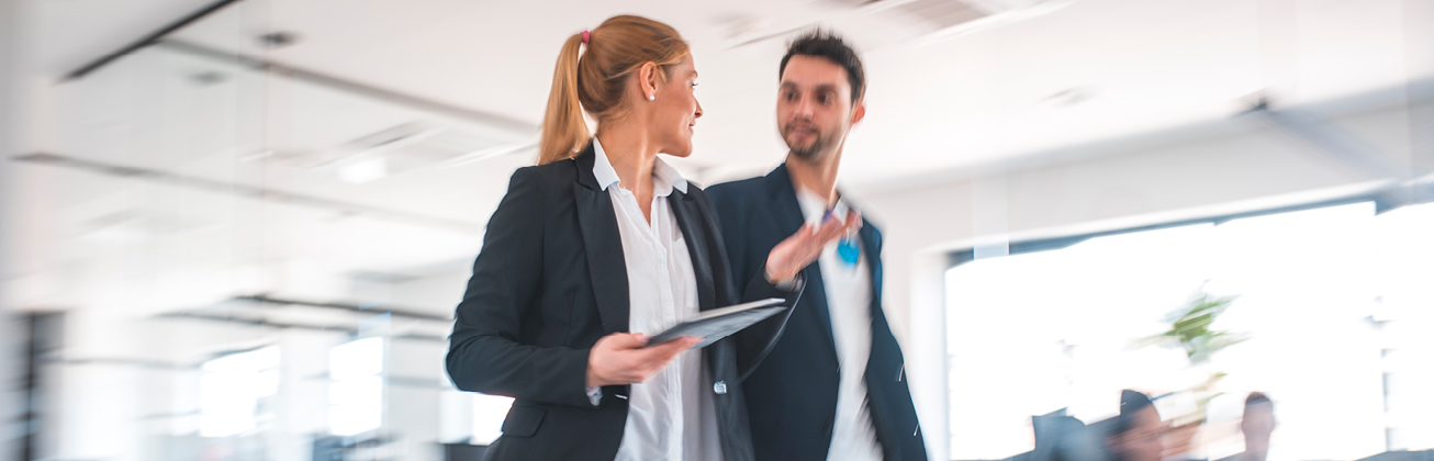 Frau mit blonden Haaren und schwarzem Blazer geht im Büro neben ihrem Kollegen mit braunen Haaren