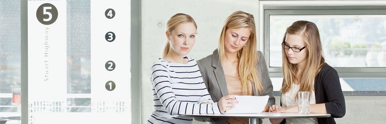 Drei blonde Studentinnen schauen sich gemeinsam an einem Hochtisch ein Dokument an