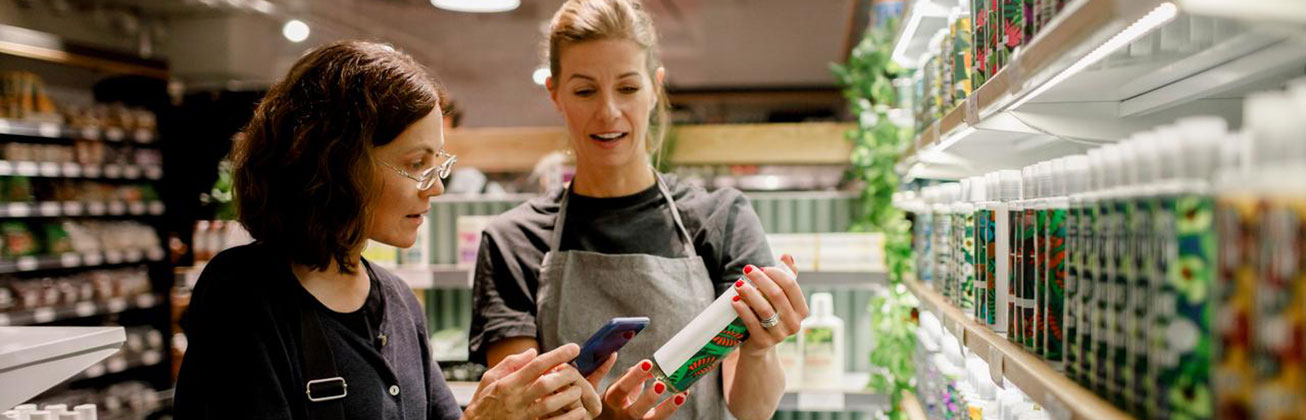 Kundin und Verkäuferin schauen vor einem Getränkeregal auf eine Flasche