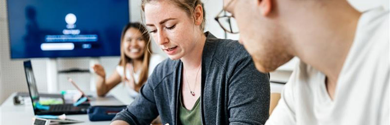 Drei Personen arbeiten gemeinsam an einem Tisch; eine Frau macht sich Notizen, während im Hintergrund eine Präsentation auf einem Bildschirm läuft.