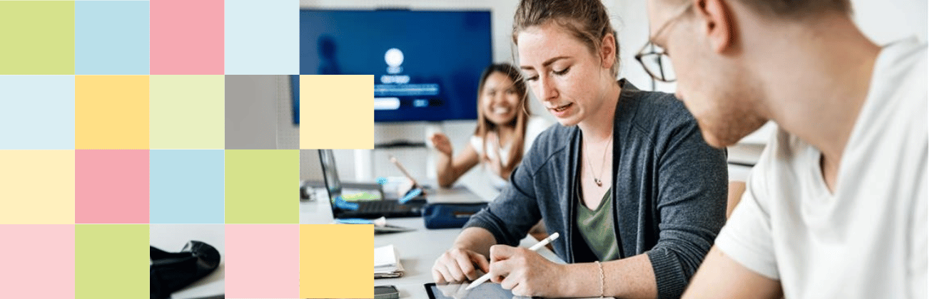 Drei Personen arbeiten gemeinsam an einem Tisch; eine Frau macht sich Notizen, während im Hintergrund eine Präsentation auf einem Bildschirm läuft.