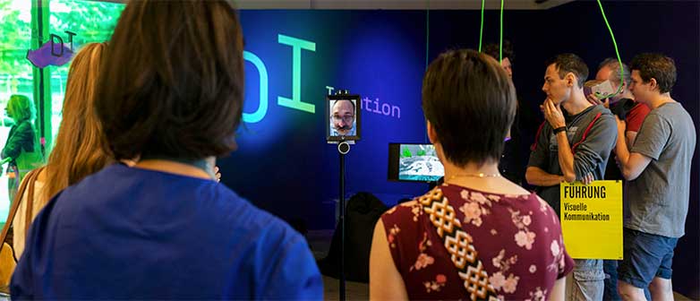Personen betrachten ein künstlerisches Werk und erhalten Informationen über eine Person im Screen
