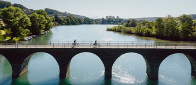     Mehrere Personen fahren mit Fahrrädern über eine steinerne Bogenbrücke über einen Fluss, umgeben von grüner Landschaft.