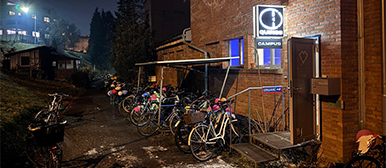 Fahrräder stehen vor einem beleuchteten Gebäudeeingang bei Nacht, mit sichtbarem Campus-Schild.