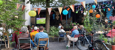    Menschen sitzen in einem begrünten Innenhof und schauen gemeinsam ein Fussballspiel auf einer Leinwand.