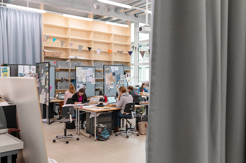     Studierende sitzen in einem hellen Atelier an Arbeitstischen und arbeiten konzentriert an Laptops und Projekten, kreatives Umfeld mit Materialien und Pinnwänden.