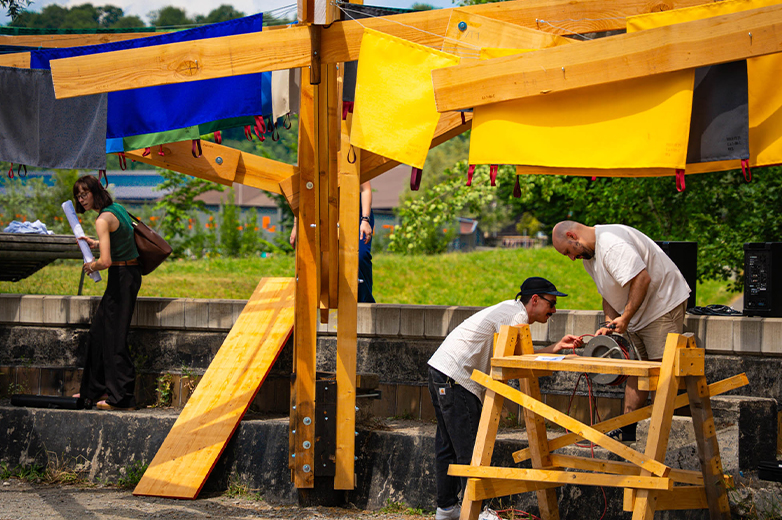     Studierende arbeiten im Freien an einer Holzkonstruktion mit Werkzeugen und farbigen Stoffen, gemeinsames handwerkliches Projekt im urbanen Raum.