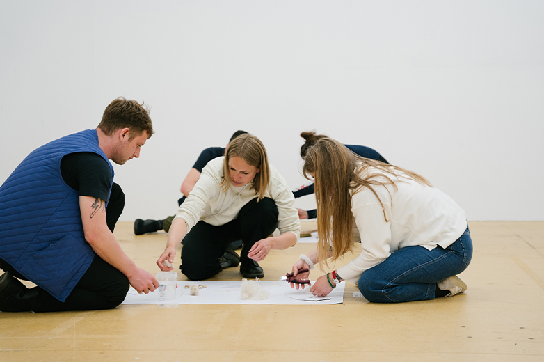     Kleine Gruppe von Studierenden kniet am Boden und entwickelt gemeinsam ein Konzept mit Materialien und Skizzen auf Papier.
