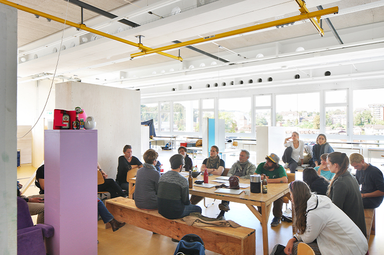     Seminar- oder Gruppensituation in einem offenen Studio, Studierende sitzen zusammen an einem grossen Tisch und diskutieren ein Projekt.