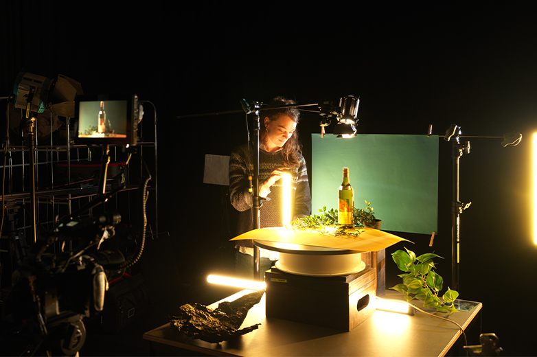     Studentin richtet ein Stillleben-Setup in einem dunklen Studio ein, mit Lichtquellen, Kamera und Objekten für eine Foto- oder Filmproduktion.