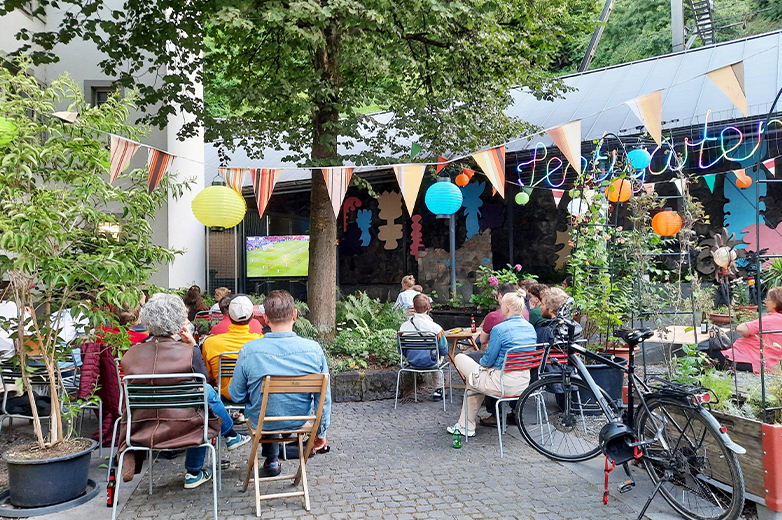     Menschen sitzen in einem begrünten Innenhof unter Lichterketten und schauen gemeinsam ein Fussballspiel auf einer Leinwand, entspannte Atmosphäre im urbanen Garten.
