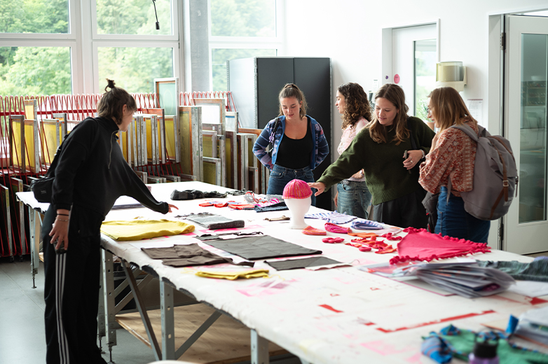     Studierende stehen um einen grossen Arbeitstisch und betrachten gemeinsam Materialproben und textile Entwürfe in einem hellen Atelierraum.