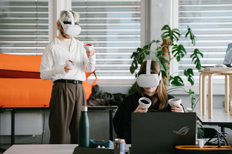     Zwei Studierende arbeiten mit Virtual-Reality-Brillen und Controllern an einem Laptop in einem modernen Arbeitsraum mit Pflanzen im Hintergrund.