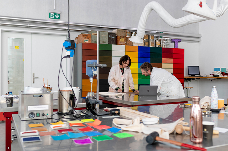     Zwei Personen arbeiten konzentriert an einem Tisch in einer Werkstatt mit bunten Materialschubladen und Werkzeugen, Fokus auf experimentelles Gestalten.