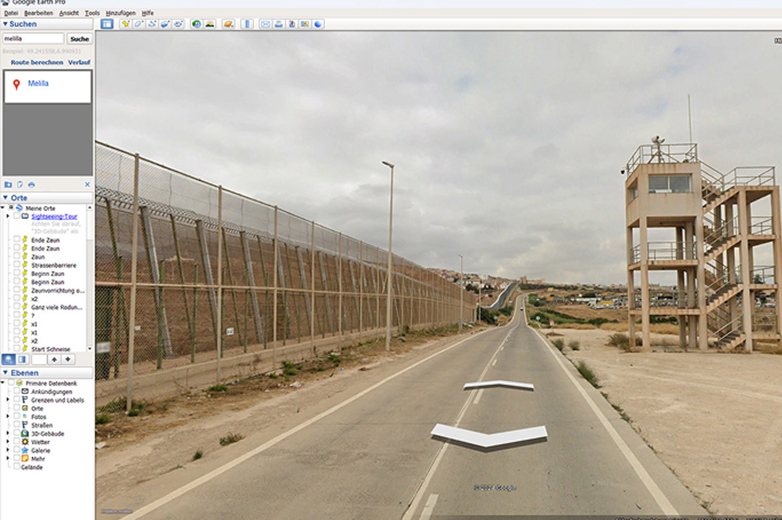 Screenshot aus Google Street View zeigt Grenzzaun bei Melilla Blick durch Street View auf eine breite Strasse mit hohen Zäunen und einem Wachturm entlang der Grenze von Melilla.
