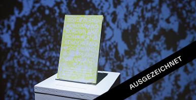 Auszeichnung für das Projekt „Borderland“ auf Podest Das Buch „Borderland“ steht auf einem Sockel, darüber ein schwarzes Banner mit dem Schriftzug „Ausgezeichnet“.