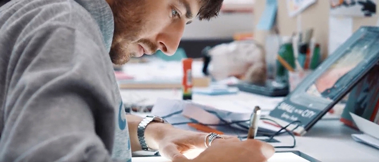 Ein Mann sitzt an einem Schreibtisch und zeichnet konzentriert auf einem Tablet; im Hintergrund sind ein Laptop und ein Pinnboard mit Notizen zu sehen.