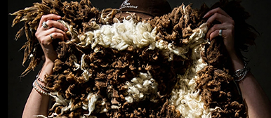     Person hält ein handgefertigtes textiles Objekt aus braunen und weissen Naturfasern vor dunklem Hintergrund.