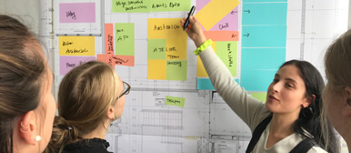 Studentin erklärt anderen Studentinnen etwas an der Wand, die mit farbigen Post-Its vollbeklebt ist