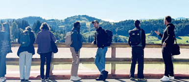 Studierende stehen in der Natur vor einem Holzzaun und blicken auf die Hügel, die sich im Hintergrund befinden