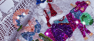     Nahaufnahme einer farbenfrohen Stickerei aus glänzenden Pailletten in Pink, Violett, Rot, Grün und Blau auf weissem Spitzenstoff.