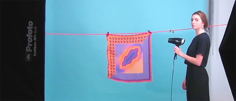 Studentin, die im Fotostudio vor einem blauen Hintergrund steht und mit einem Föhn ein farbiges Seidentuch anbläst