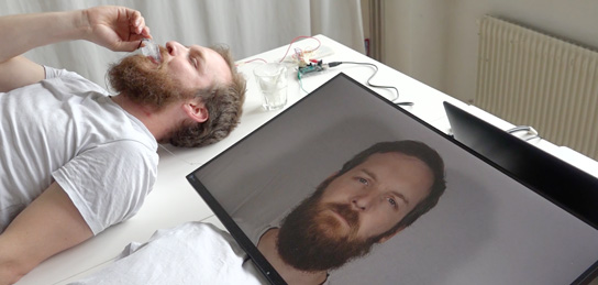 Ein Typ liegt auf einem Bett. Neben ihm ein Bildschirm, der eine Ansicht seines Kopfes zeigt.