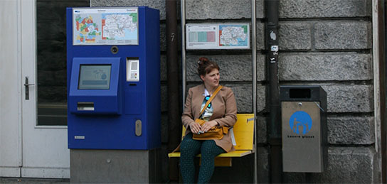 Frau sitzt auf einem Klappbänkli an einer Bushaltestelle