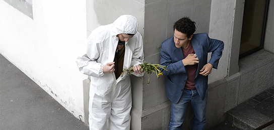 Zwei junge Männer treffen sich an Hausecke, der rechts in weissem Overall mit Blumen in der Hand der Links in blauem Anzug